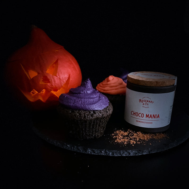 Ein geschnitzter Kürbis, zwei Muffins mit lila und oranger Glasur und eine Dose auf einem schwarzen Teller.