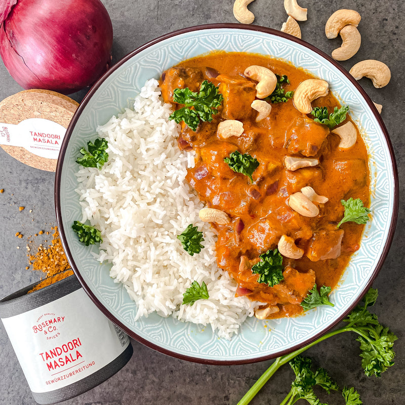 Ein Teller mit Reis und Tandoori-Curry garniert mit Cashewnüssen und Petersilie, daneben eine Dose Tandoori Masala.