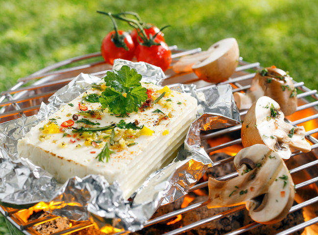 Fetakäse mit Kräutern und Gewürzen auf Alufolie, grilliert mit Pilzen und Tomaten, im Hintergrund grüne Wiese.
