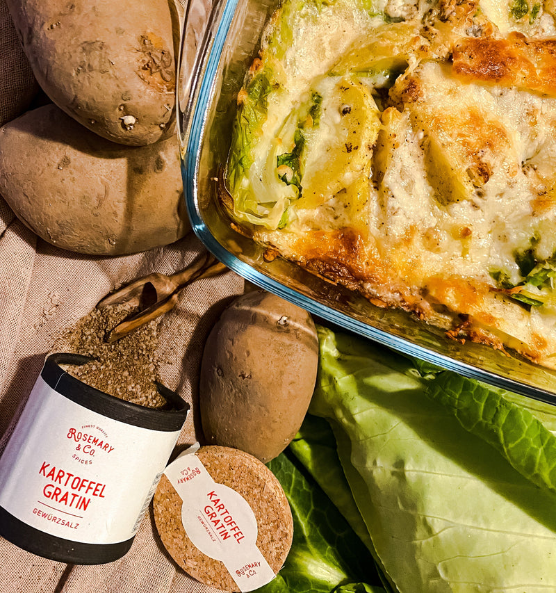Kartoffelgratin in einer Glasform, umgeben von frischen Kartoffeln, Kohl und einer Dose mit Kartoffelgratin-Gewürz.