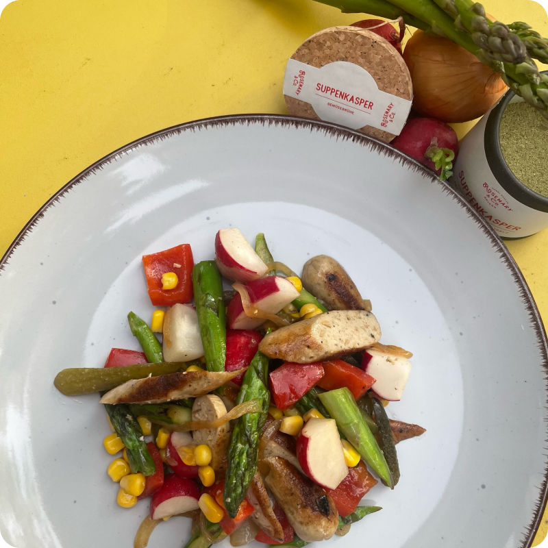 Ein Teller mit Gemüse und Wurststücken, im Hintergrund eine Dose und einige frische Zutaten auf einem gelben Tisch.