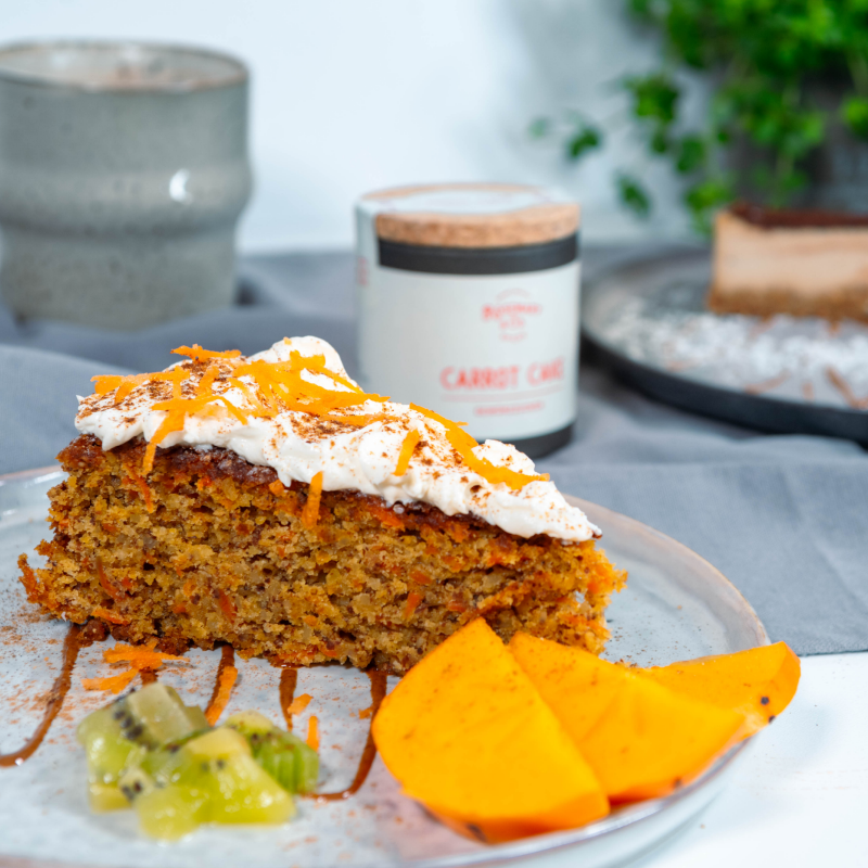 Ein Stück Karottenkuchen mit Sahne und geriebener Karotte auf einem Teller mit Kiwi- und Mangostücken serviert.