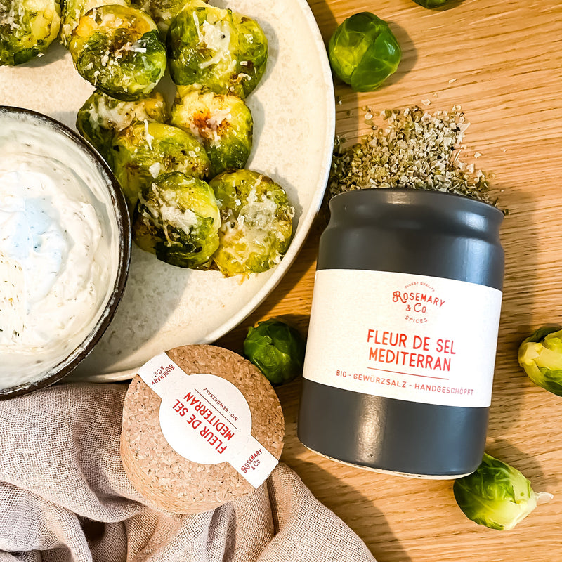 Ein Teller mit geröstetem Rosenkohl, einer Schale Dip und einer dunklen Dose mit Fleur de Sel auf einem Holztisch.