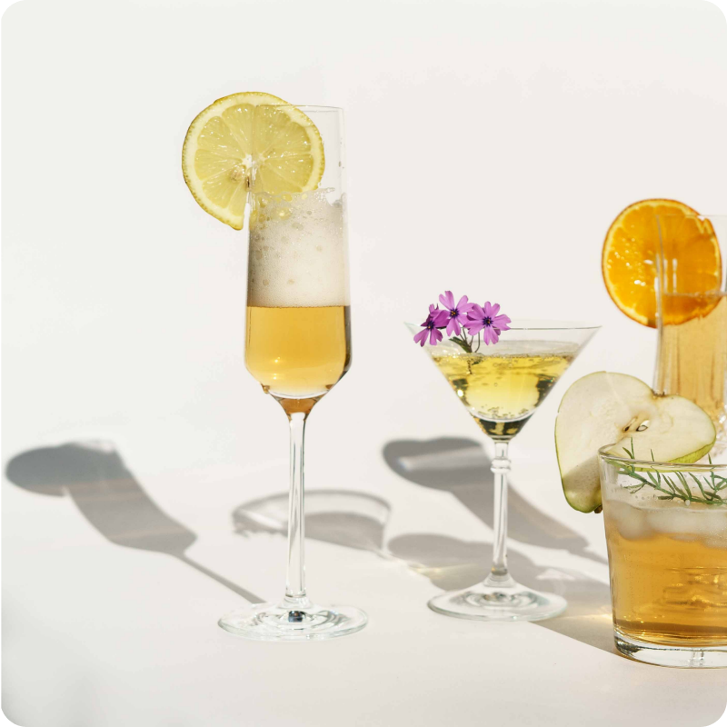 Verschiedene elegante Gläser mit Getränken, garniert mit Zitrus- und Apfelscheiben sowie Blumenakzenten.