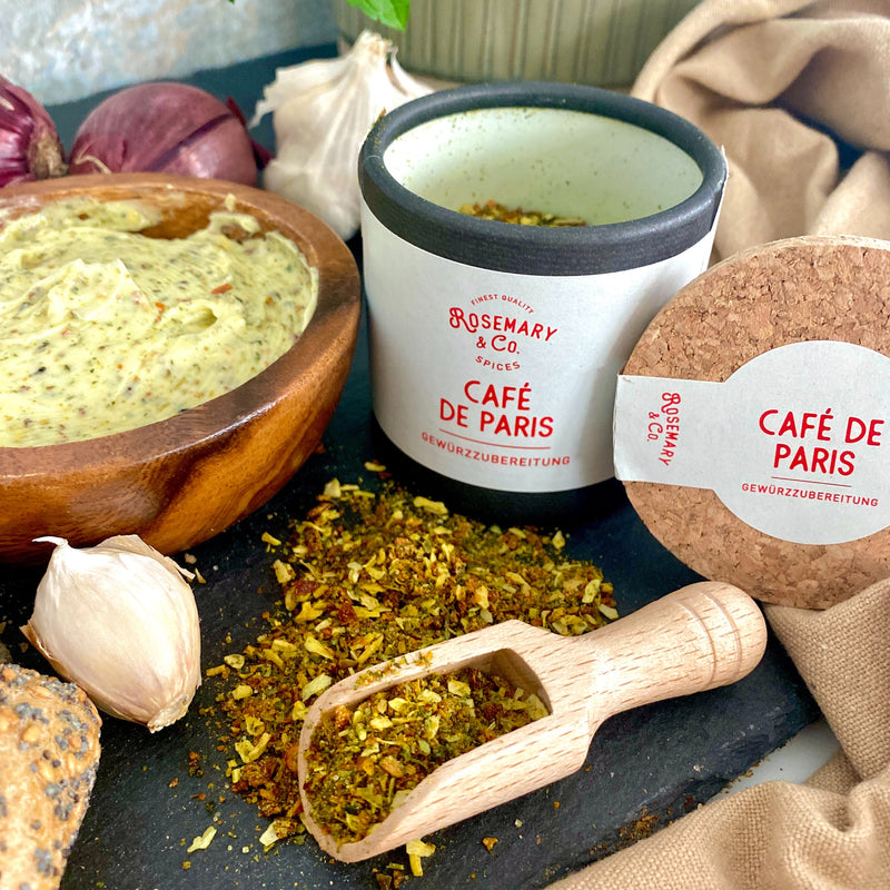 Eine Pappdose mit "Café de Paris" Gewürz und Gewürzmischung auf einem Tisch mit Knoblauch, Zwiebel und Kräuterbutter.
