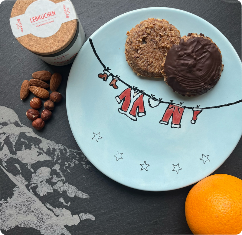 Ein Teller mit zwei Lebkuchen, daneben eine Dose mit Korkdeckel, Mandeln, Haselnüssen und eine Orange.