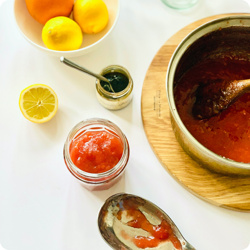 Ein Topf mit roter Marmelade, ein Glas mit Marmelade, ein Teller Zitronen und ein Löffel auf einem weißen Tisch.