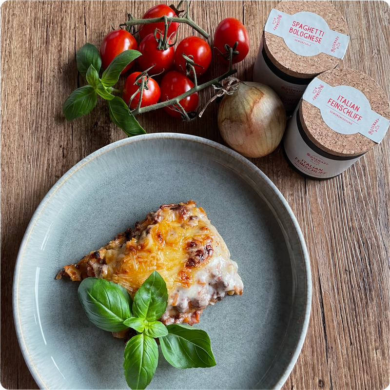 Ein Teller Lasagne mit Basilikum, Tomaten, Zwiebel und zwei Pappdosen Gewürzen auf einem Holztisch.