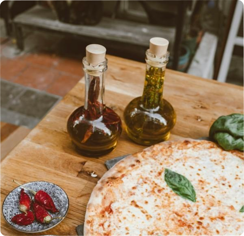 Pizza mit Basilikum, zwei Ölflaschen und Chilischoten auf einem Holztisch.