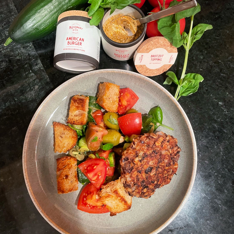 Ein Teller mit Tomatensalat, Brotstücken und einem vegetarischen Patty, daneben Gewürzdosen und frische Zutaten.