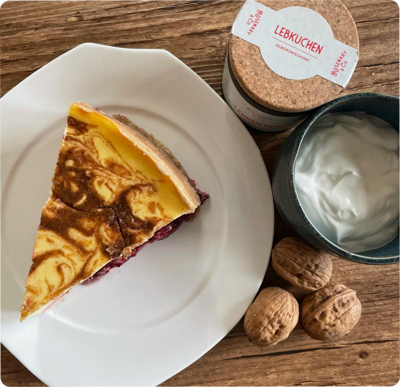 Ein Stück Kuchen auf einem Teller neben einer Schale Joghurt, drei Walnüssen und einem Gewürzglas auf einem Holztisch.