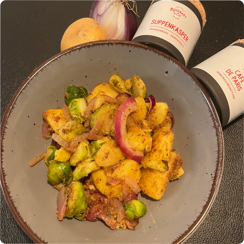 Grauer Teller mit Kartoffeln, Rosenkohl und roten Zwiebeln; daneben Gewürzdosen und frisches Gemüse.