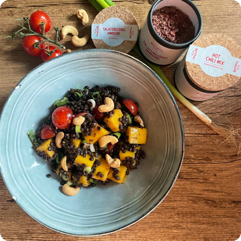 Salat in einer Schüssel mit Linsen, Kirschtomaten, Kürbis und Cashewkernen; daneben zwei Dosen mit Gewürzen.
