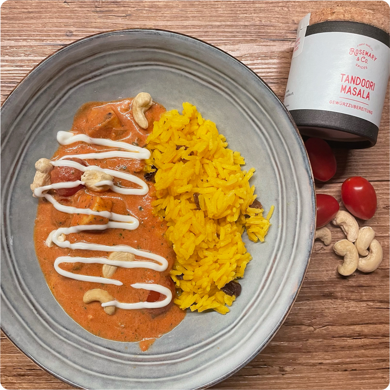 Grauer Teller mit Tandoori Masala, Cashewkernen und gelbem Reis, daneben eine Gewürzdose, Tomaten und Cashewkerne.