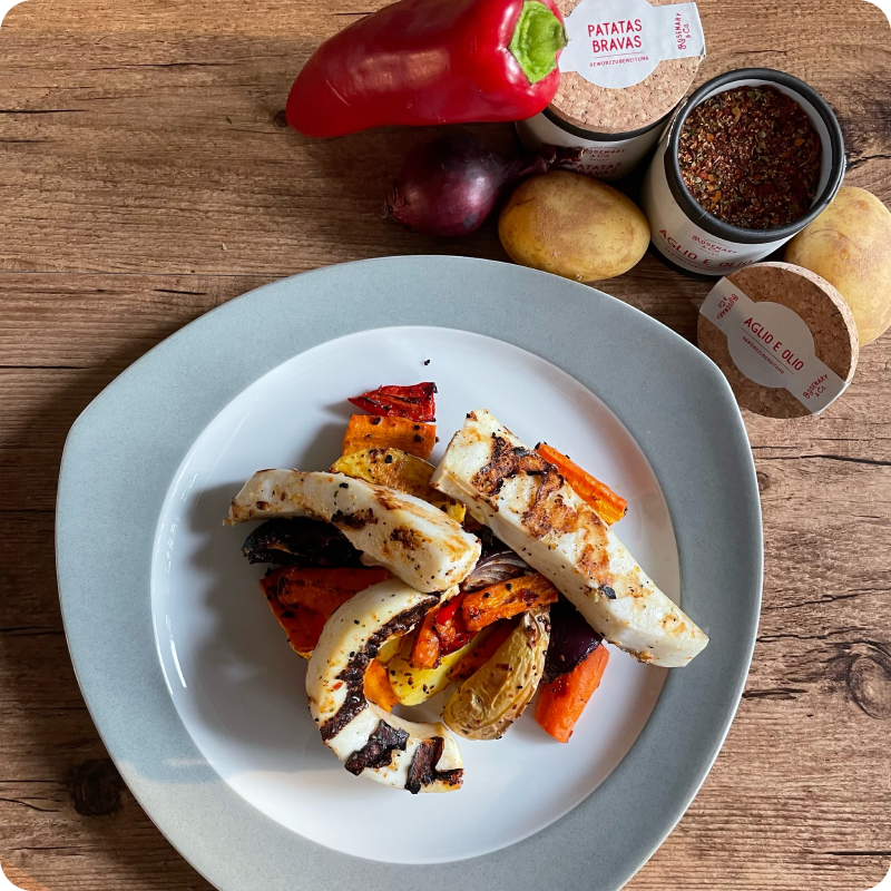 Gegrilltes Gemüse mit Halloumi auf einem Teller, daneben Paprika, Zwiebel, Gewürze in Pappdosen und Kartoffeln auf Holztisch.