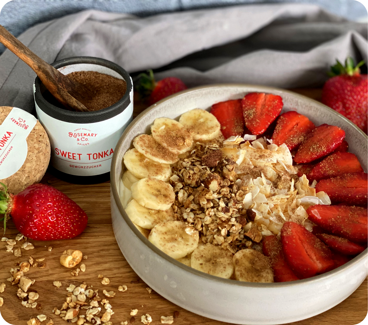 Schale mit Müsli, Banane, Erdbeeren, Kokosflocken und braunem Zucker neben Dose Sweet Tonka von Rosemary & Co