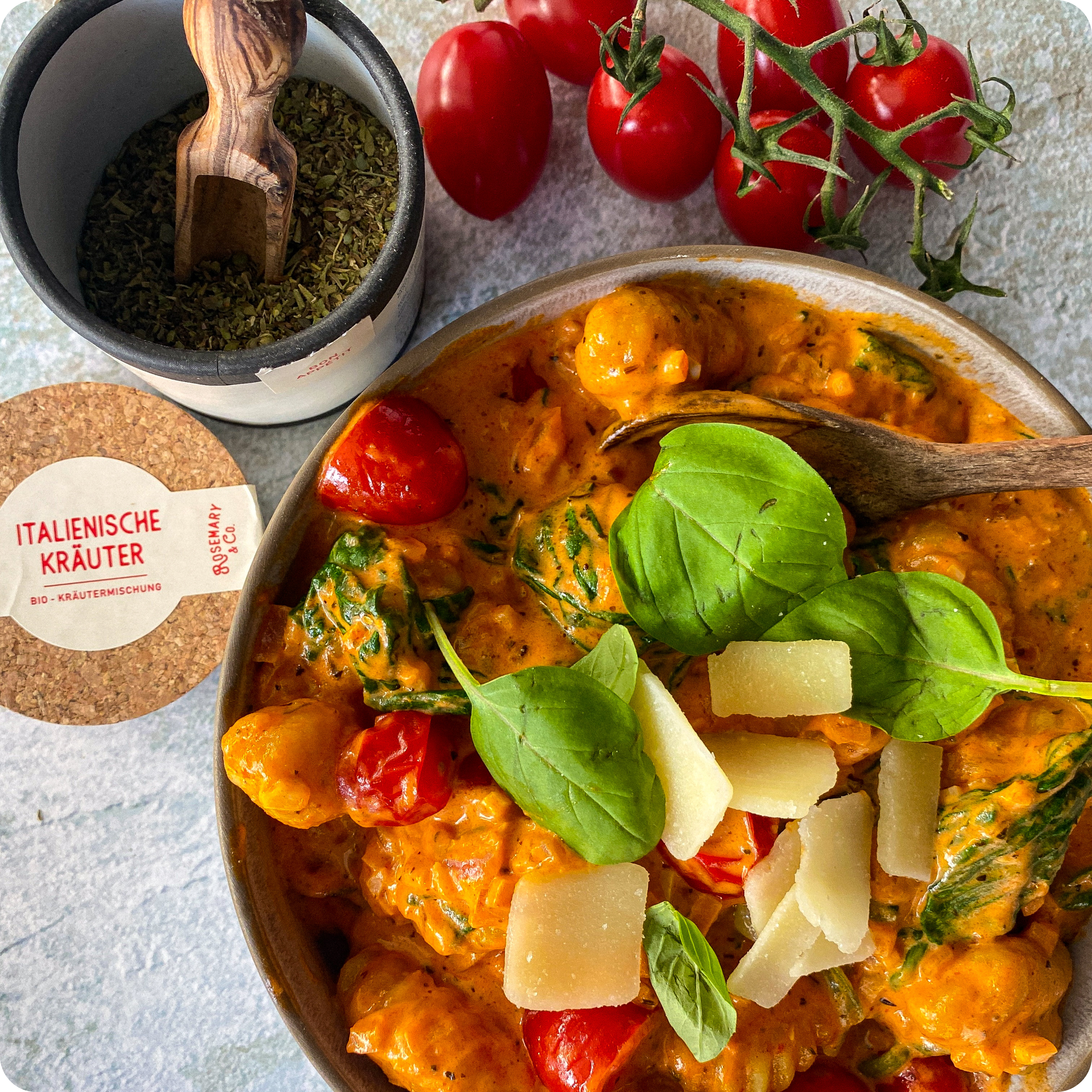 Eine Schüssel mit Tomaten-Gnocchi in cremiger Sauce, garniert mit Basilikum und Parmesan, daneben italienische Kräuter.