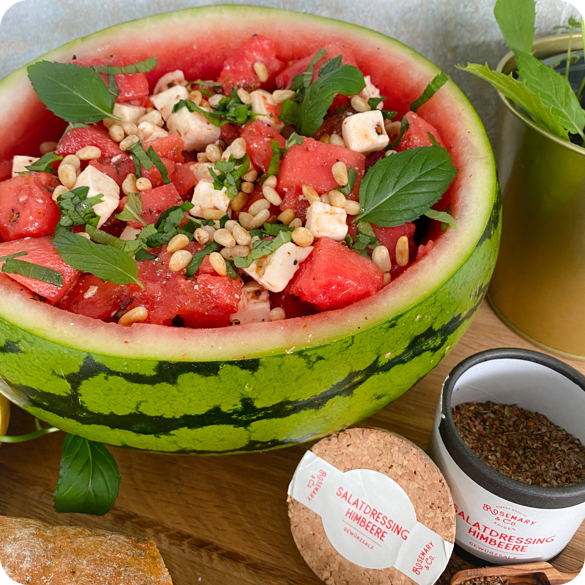 Halbierte Wassermelone als Schale mit Wassermelone, Feta, Kräutern, Pinienkernen, Dose Rosmarin & Co. Salatdressing Himbeere