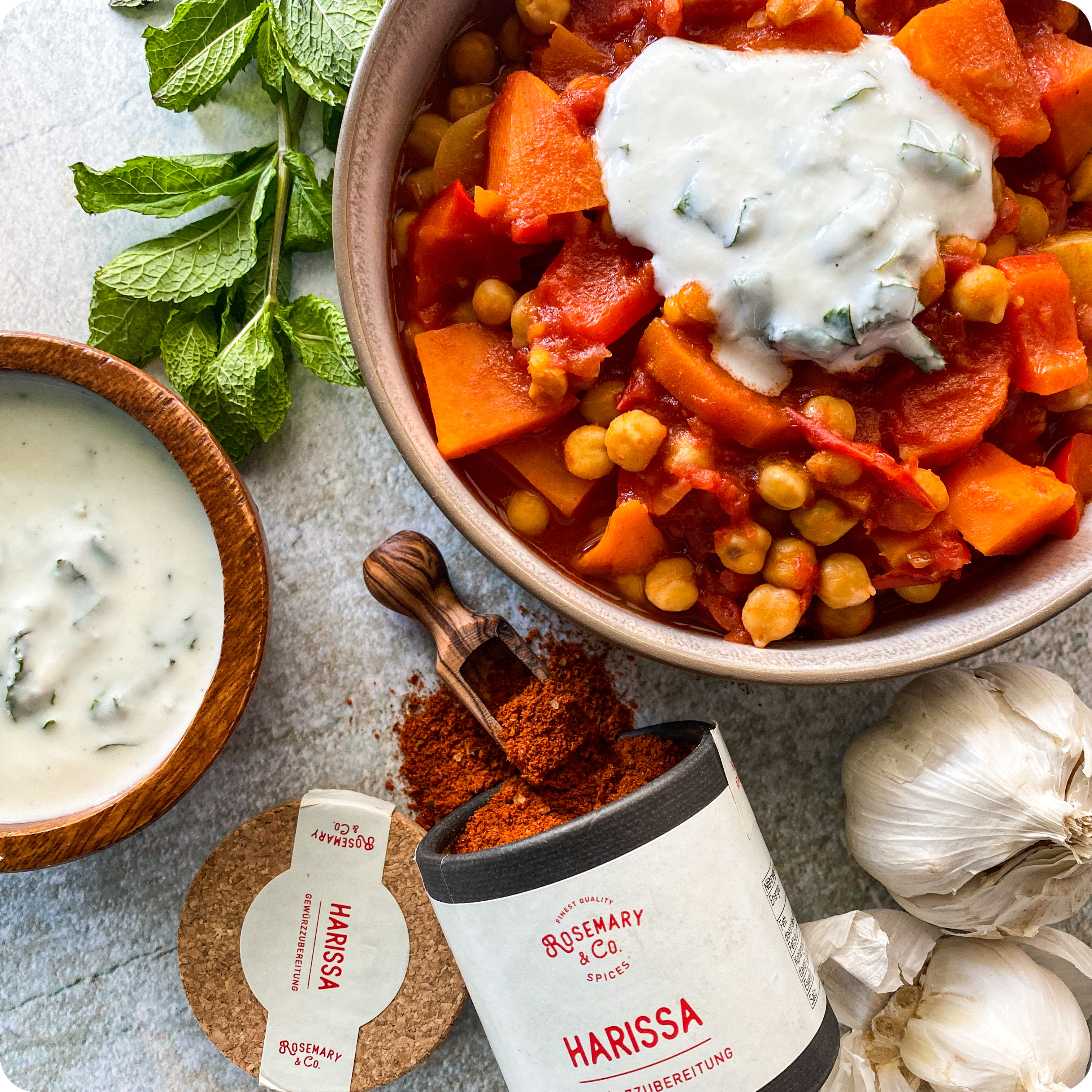 Kichererbseneintopf mit Tomaten, Karotten und Joghurt in einer Schale, dazu Dose Rosemary & Co. Harissa und frische Minze