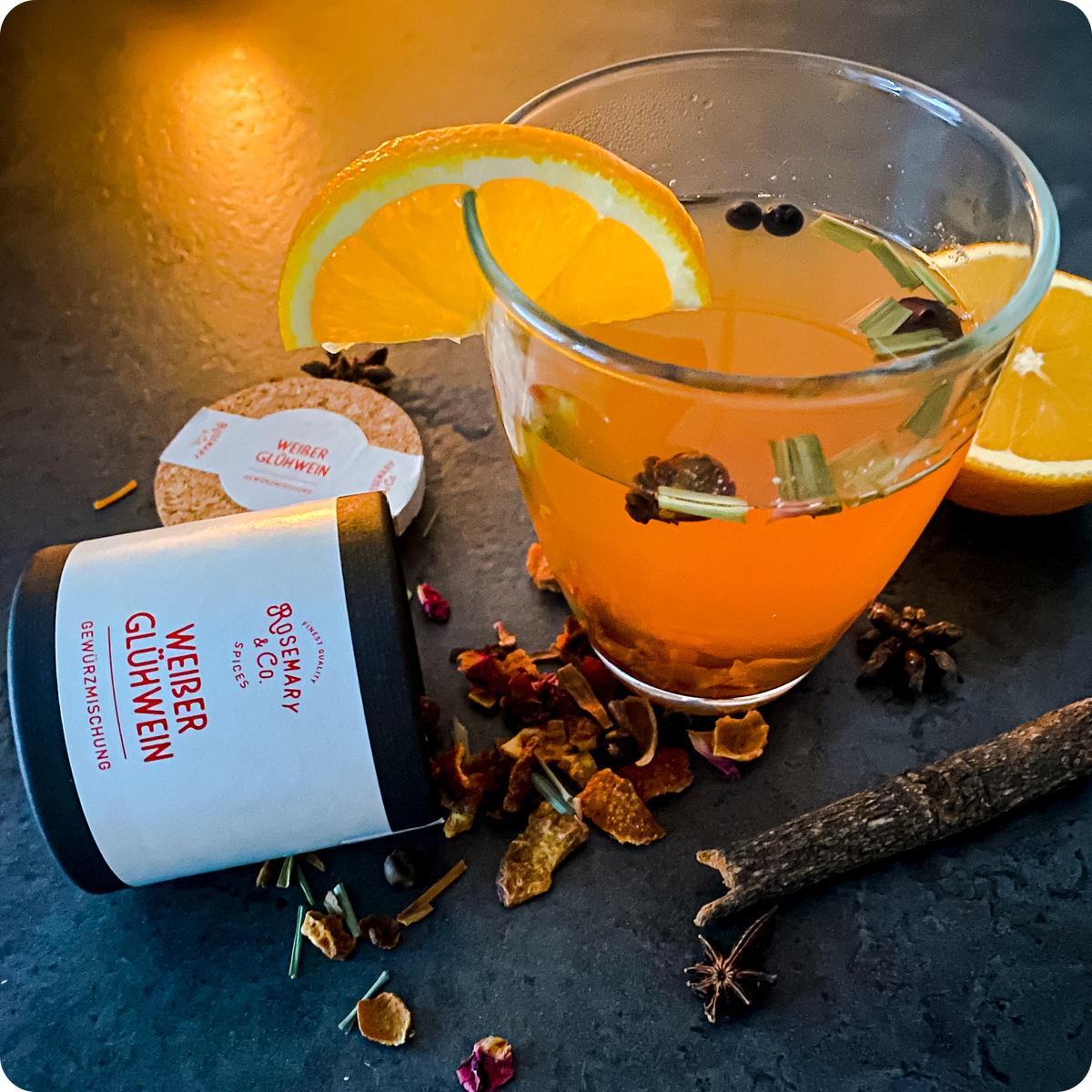 Dose orangefarbenes Getränk mit Gewürzen und Orangenscheiben neben Rosemary & Co. Gewürze Weißer Glühwein auf dunkler Fläche