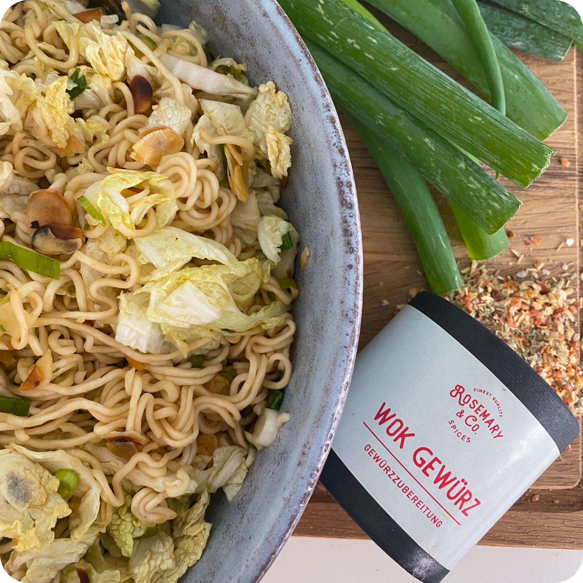 Schüssel mit Nudelpfanne, Kohl und Frühlingszwiebeln, daneben frische Frühlingszwiebeln und Dose Rosemary & Co. Wok Gewürze