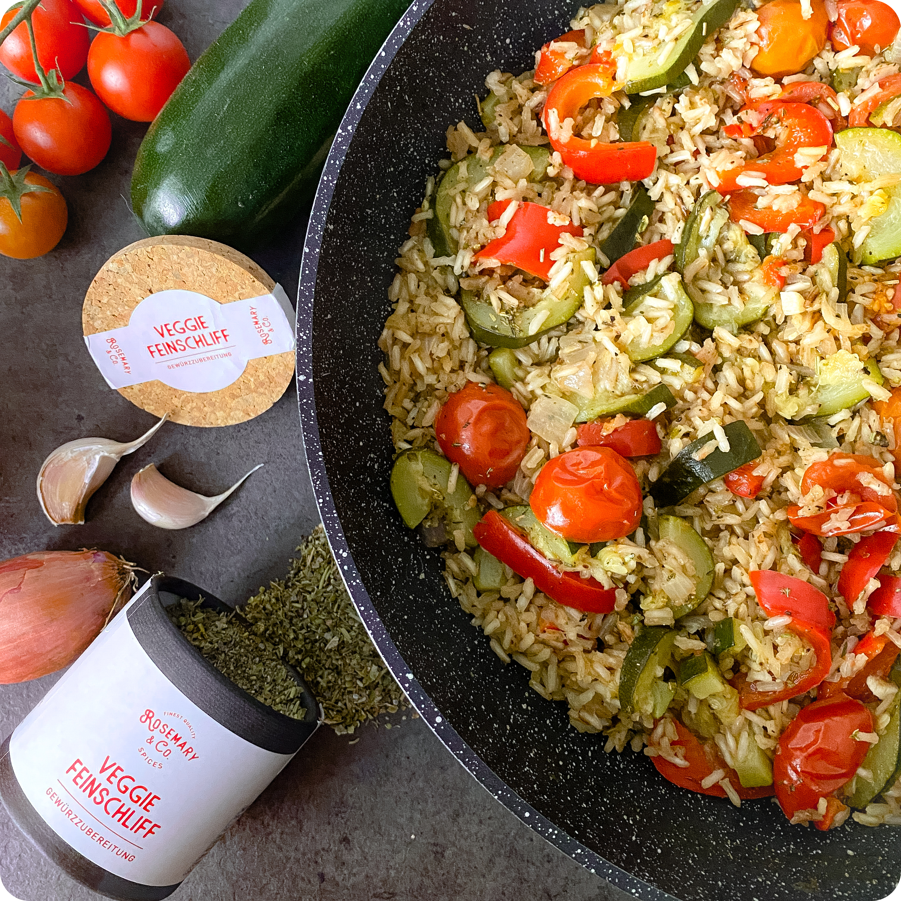 Reispfanne mit Zucchini und Paprika in der Pfanne, umgeben von Gewürzdose, Knoblauch, Tomaten und Zwiebel.