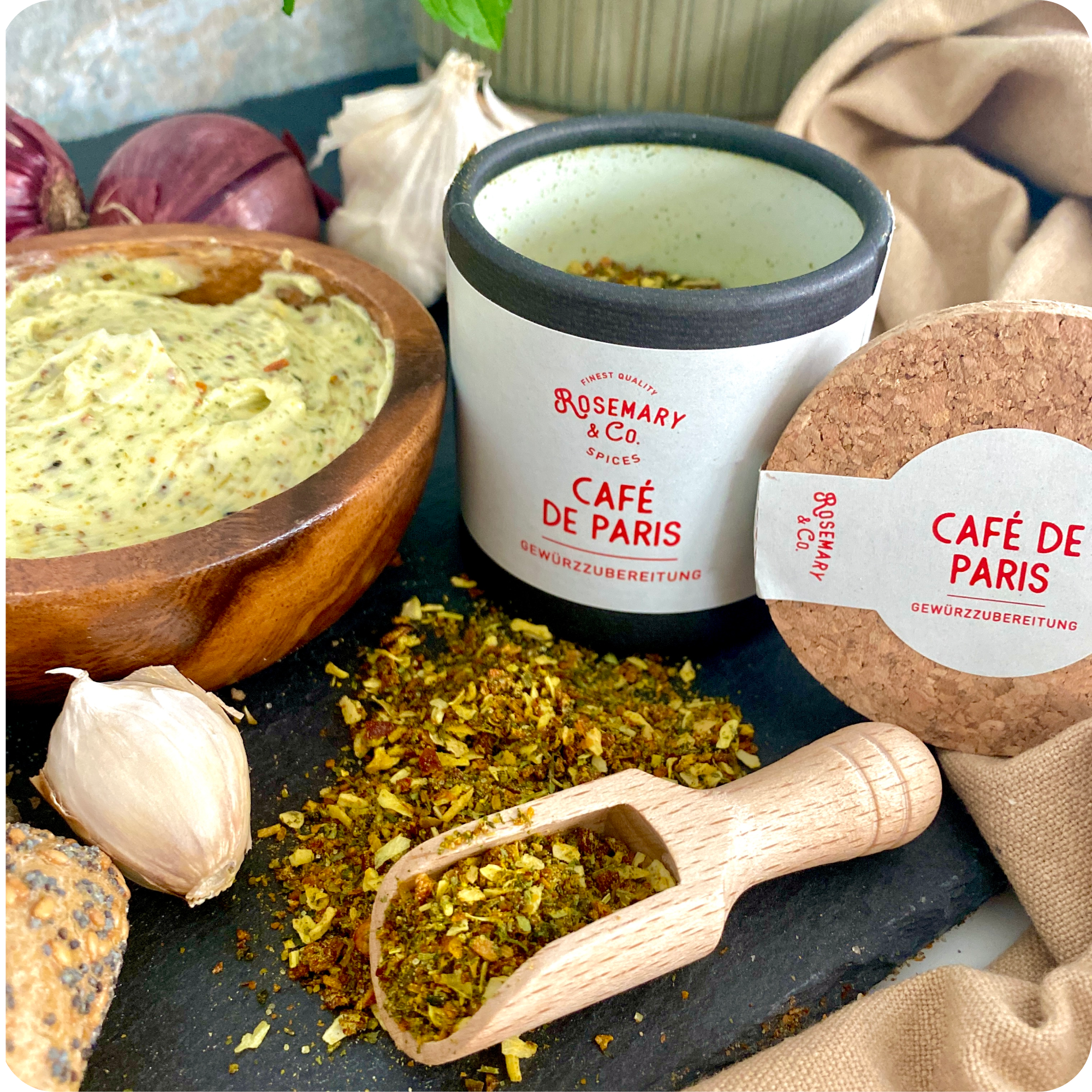 Dosieren Sie Rosemary & Co. Gewürze Cafe de Paris neben Holzlöffel, Schälchensauce, Knoblauch und Zwiebeln